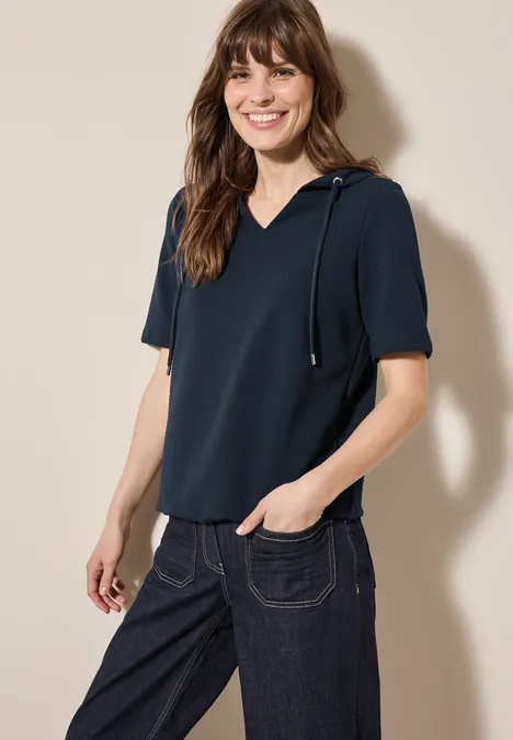 Frau mit dunkelblauem Shirt und Jeans lächelt vor hellem Hintergrund.